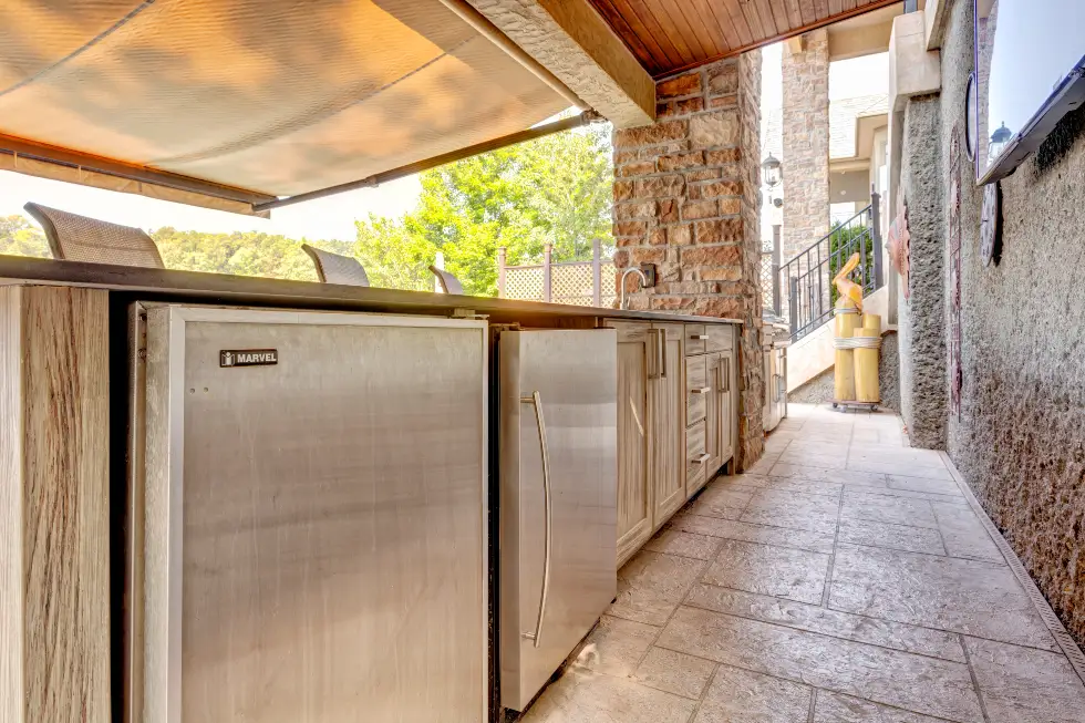 Custom covered outdoor kitchen with light wood cabinetry, stainless appliances, beverage refrigeration and stone countertops by Ragan Cabinets.